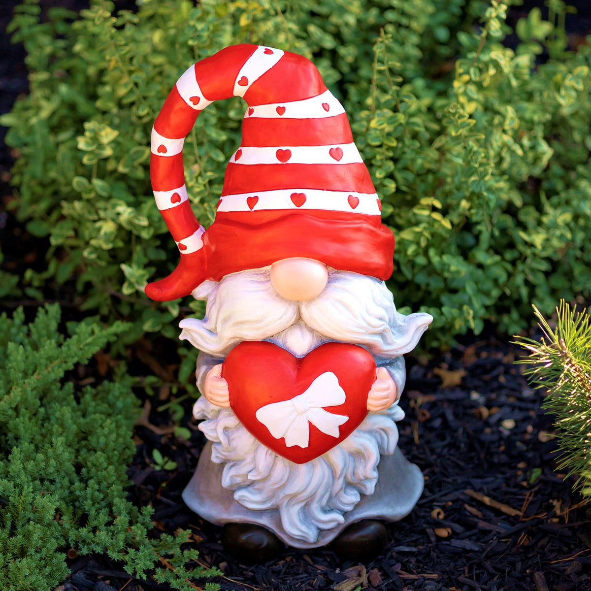Valentine gnome holding a red heart, front view among green garden plants
