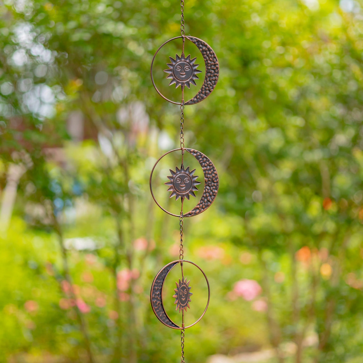 Antique bronze sun and crescent moon rain chain hanging among green garden foliage