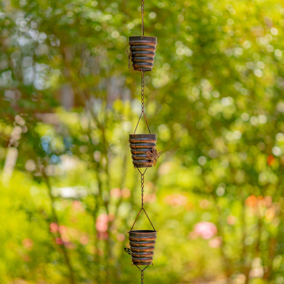 Antique bronze rain chain with bucket hive cups hanging against a green garden backdrop