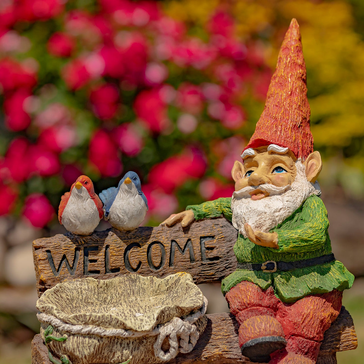 A seated gnome beside a birdbath and two birds perched on a welcome sign, with pink flowers behind.