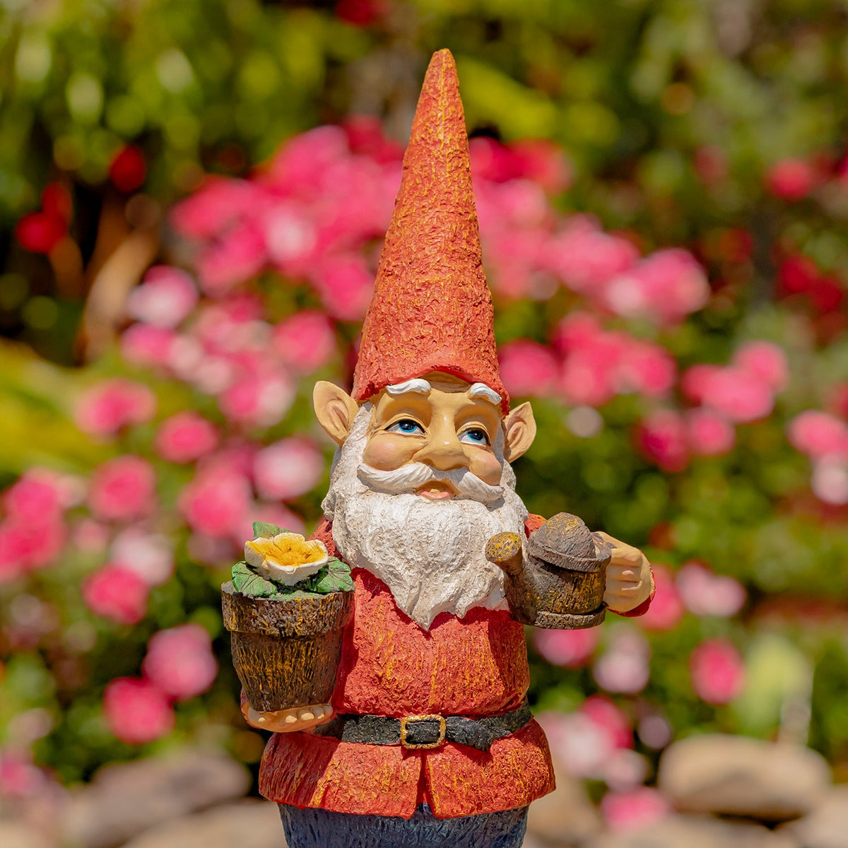 Red-hatted gnome holds a flower pot and watering can in front of pink garden blooms.