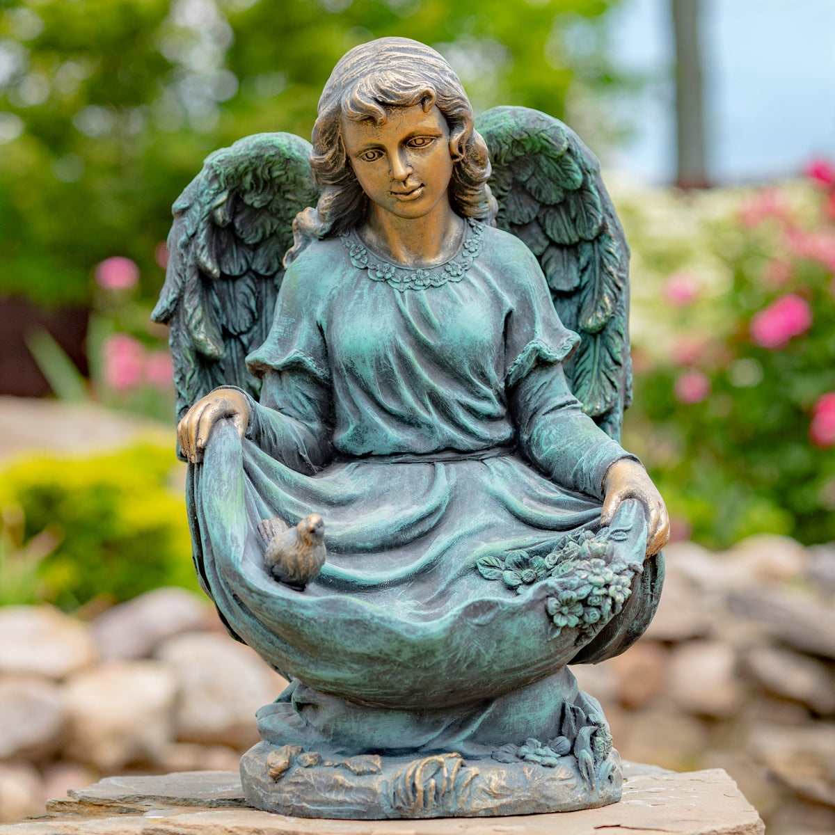 Kneeling angel statue with a bird in its draped skirt, shown on a stone surface in a garden.