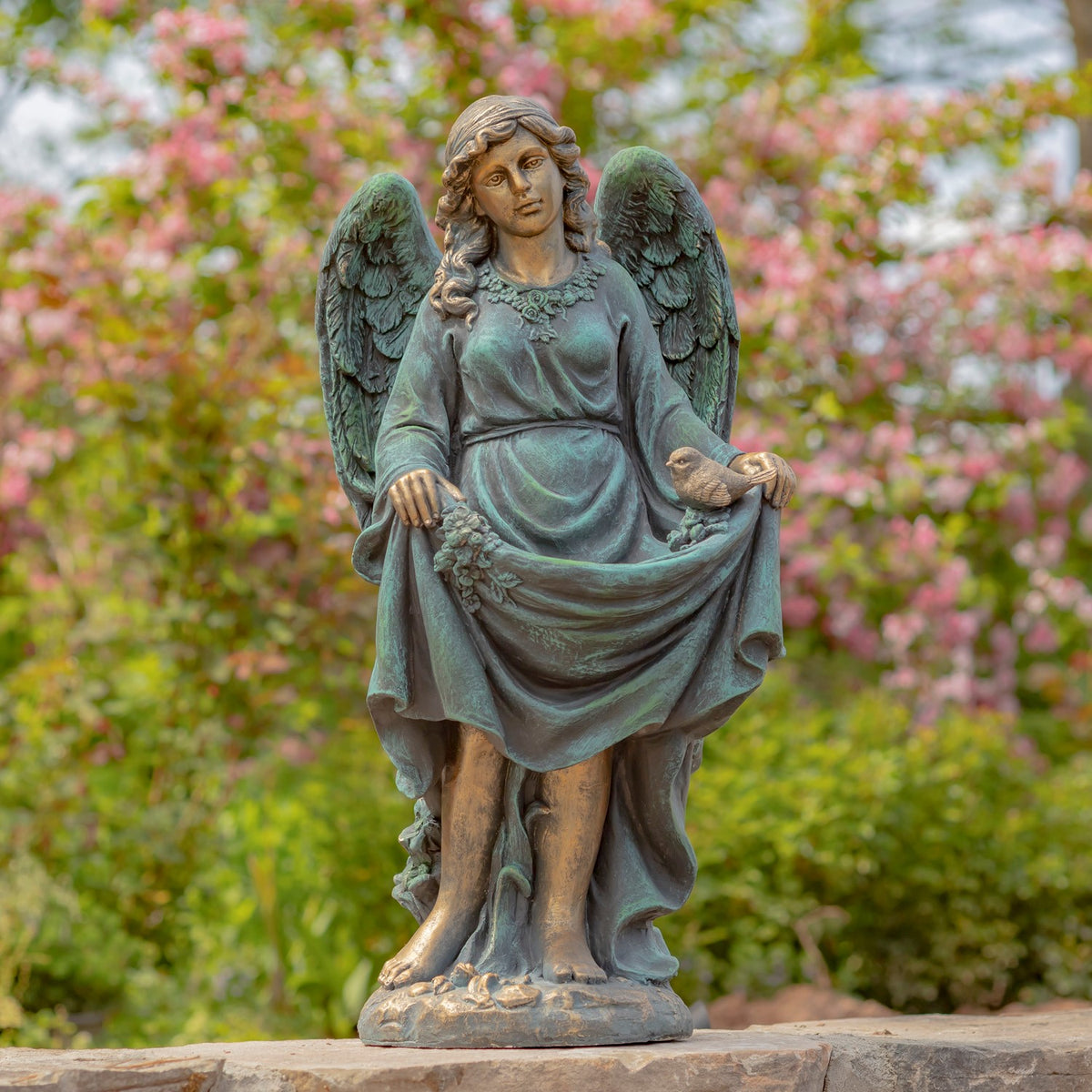 Full view of a winged statue holding out her dress, standing on a stone base among flowering shrubs.
