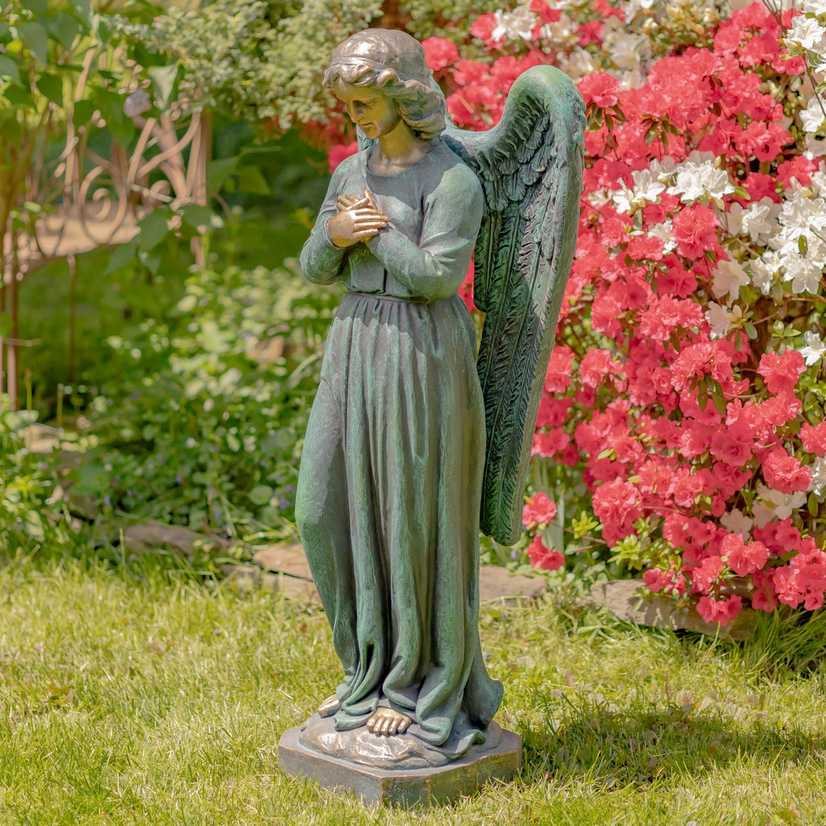 Full view of a robed angel statue with large wings standing on a base among garden flowers.