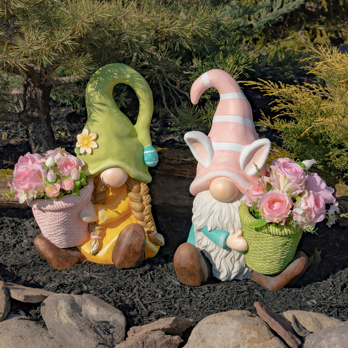 Two seated garden gnomes with pink flower-filled planters sit among mulch and rocks.
