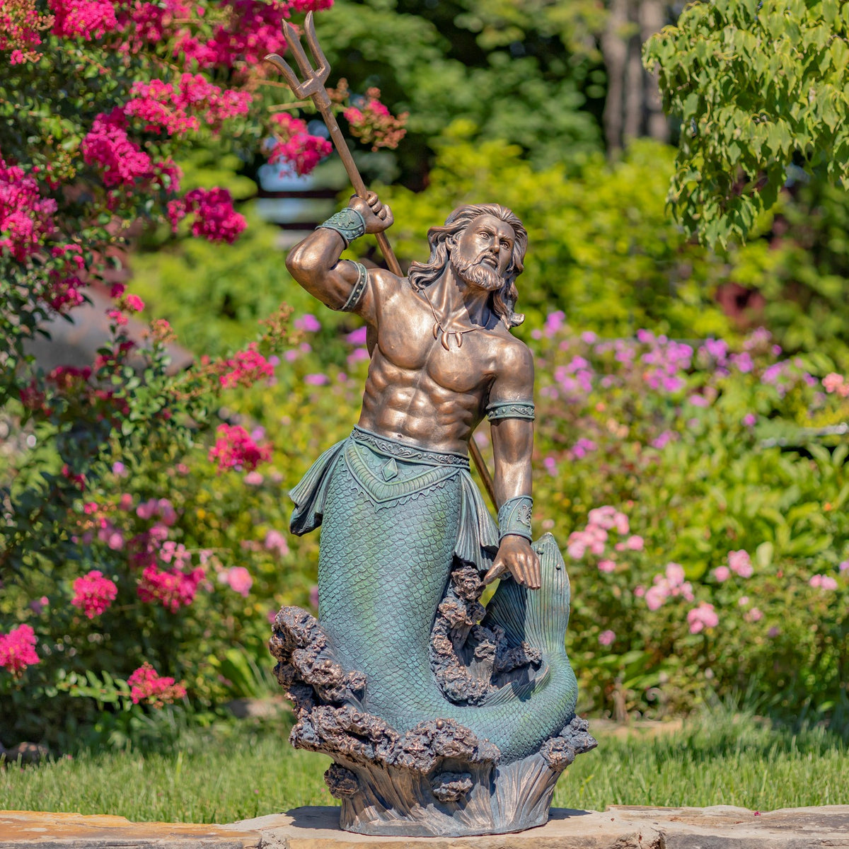 A bronze-toned merman statue raises a trident among flowering shrubs and greenery.