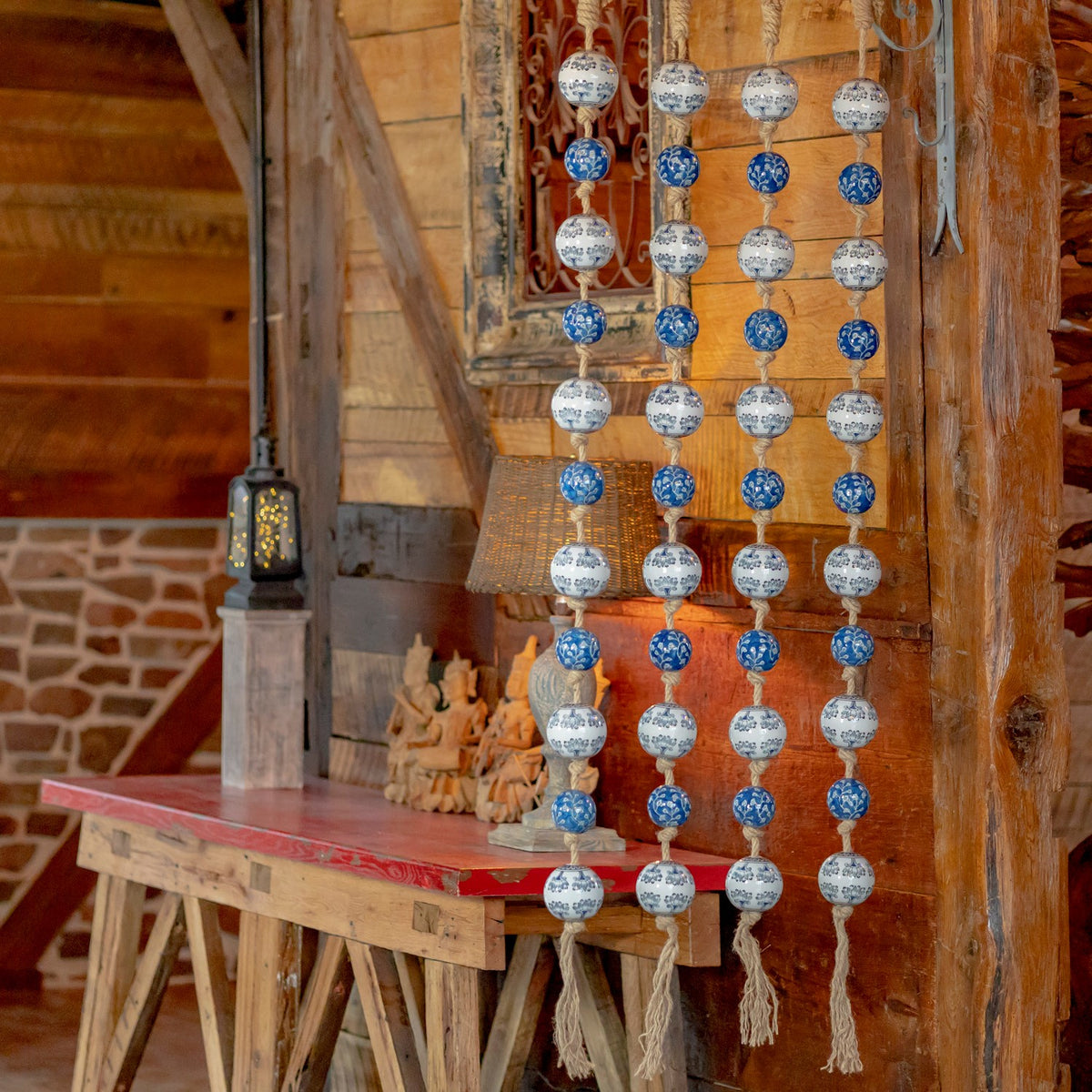 Four strands of blue and white patterned ceramic balls hang against a rustic wood wall beside a table.