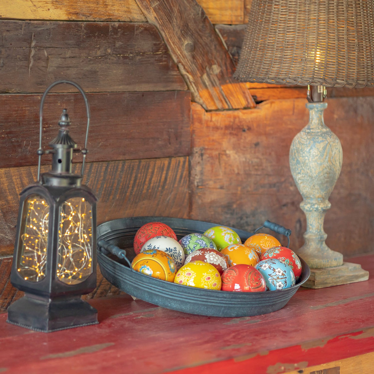 A shallow dark metal bowl filled with colorful patterned ceramic balls sits between a lantern and a table lamp.