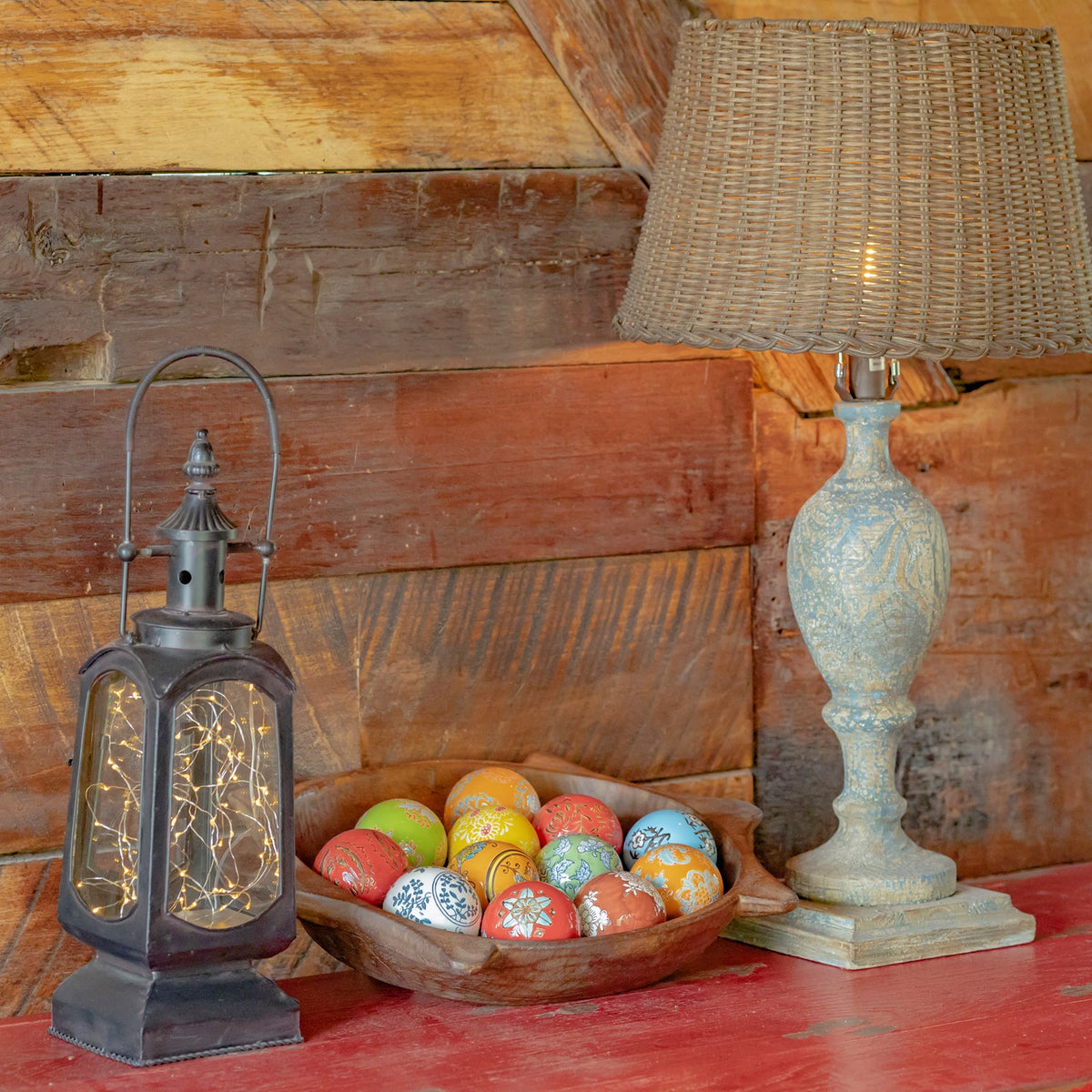 A bowl of colorful patterned ceramic balls sits on a red tabletop between a lantern and a lamp.
