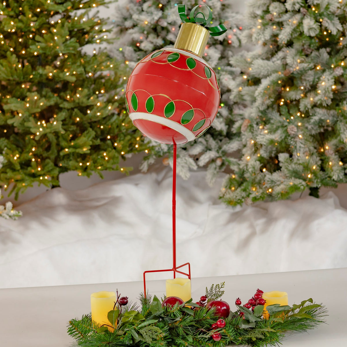 Red ball ornament garden stake displayed among a Christmas wreath and candles beneath snowy evergreen branches.