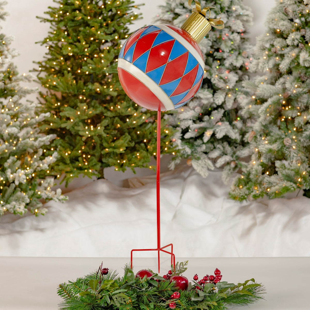 Red and blue diamond-patterned ornament on a tall red stake, displayed among greenery and Christmas trees.