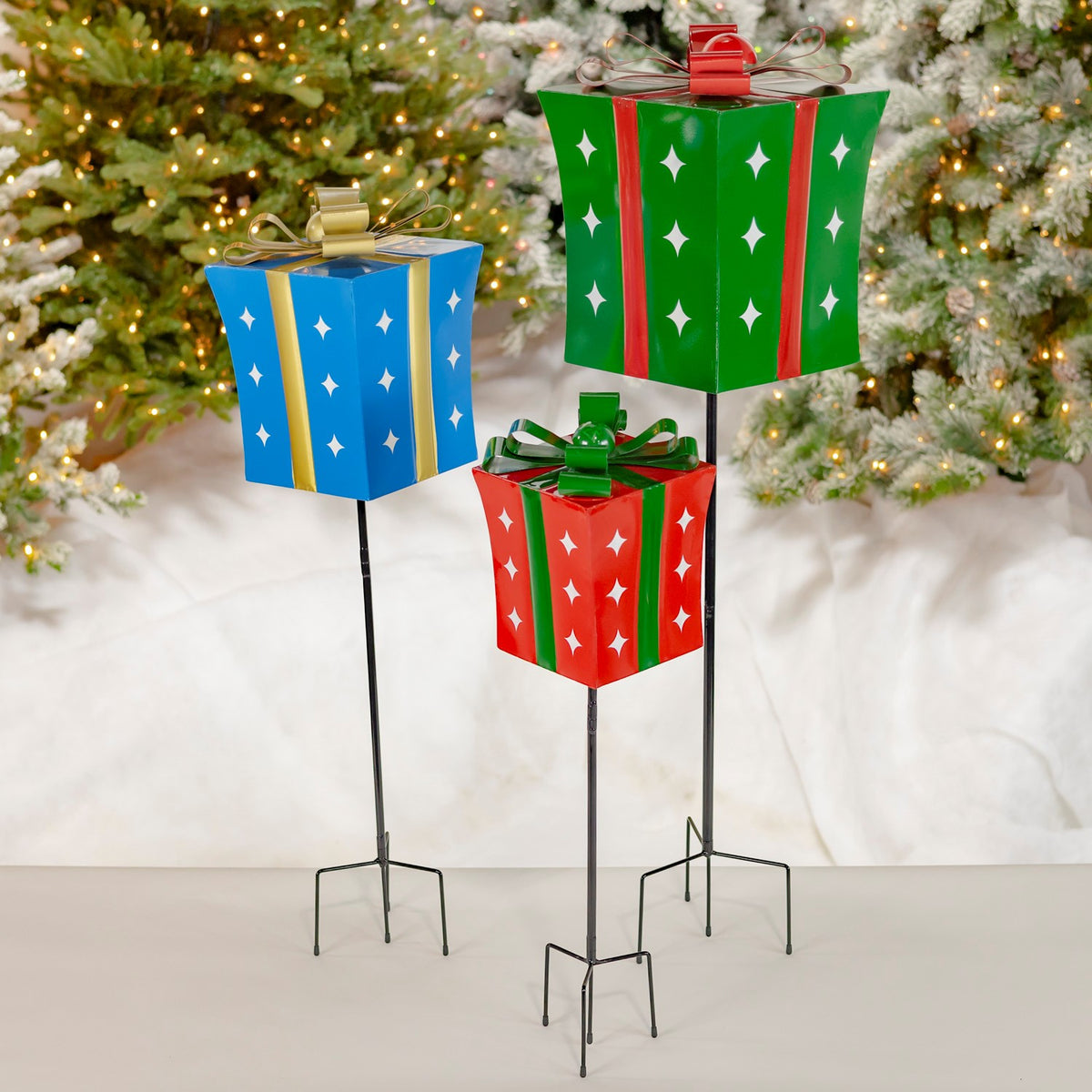 Three colorful gift-box garden stakes stand in snow beneath a lit Christmas tree.