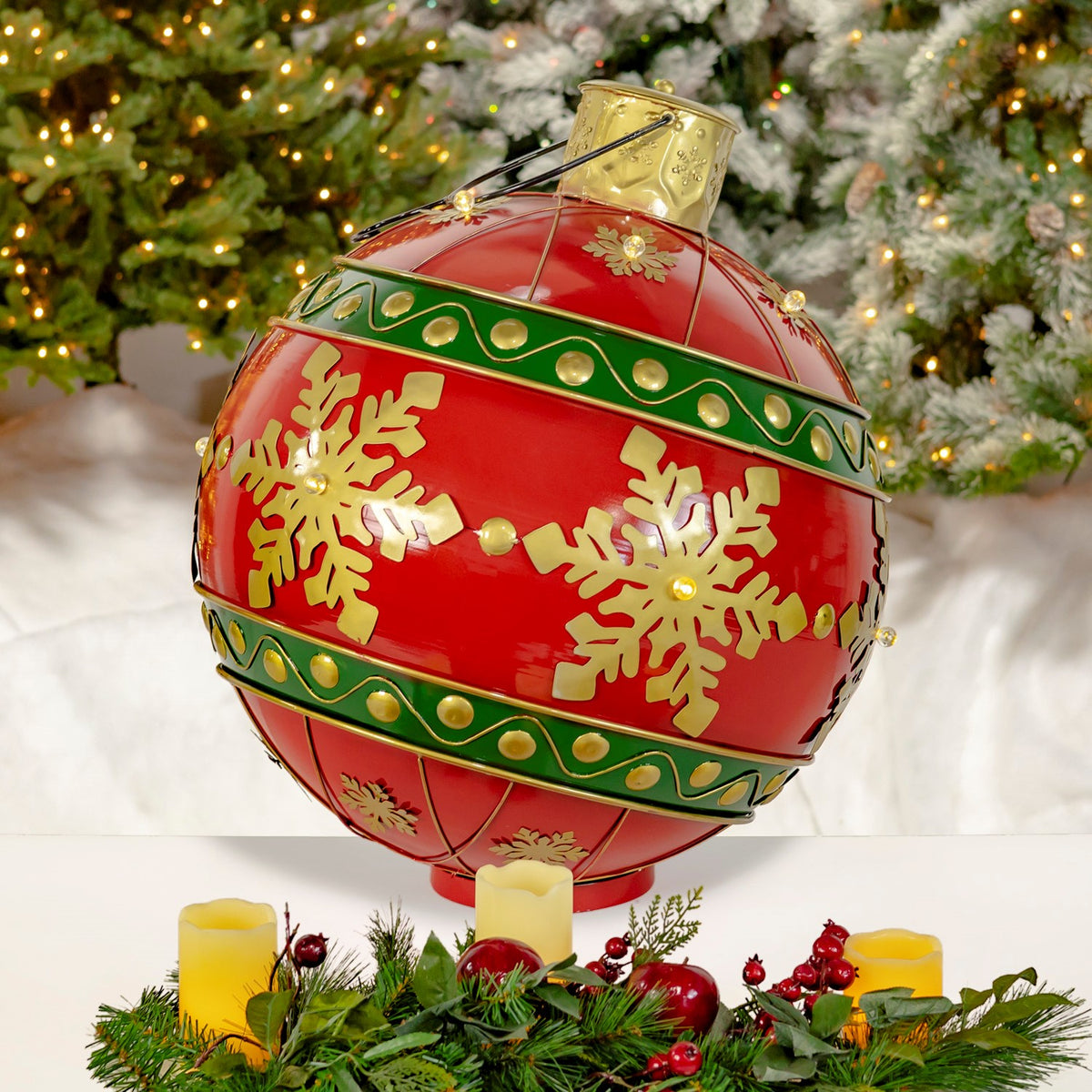 Large red Christmas ball with gold snowflakes and green bands displayed among candles and evergreen branches.