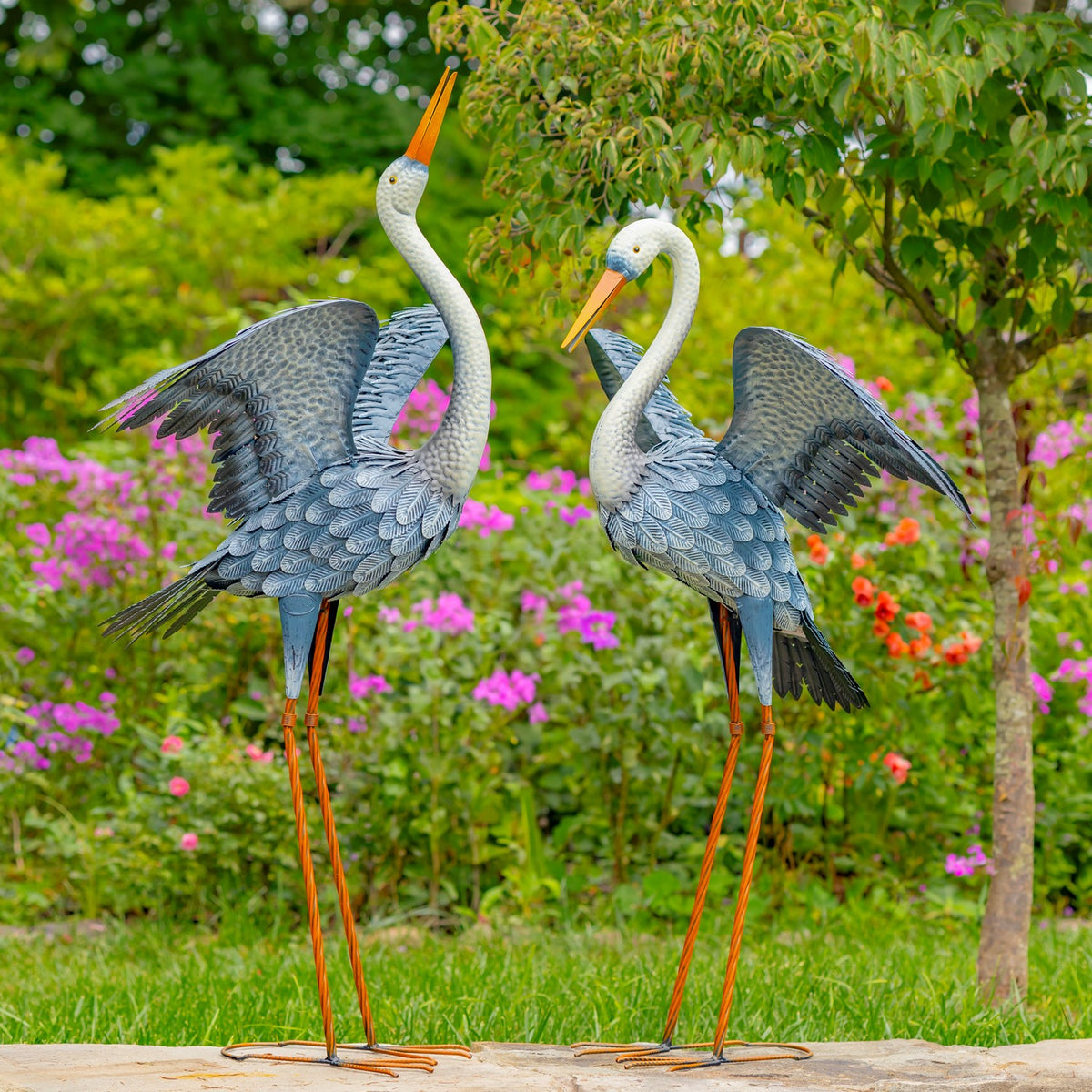 Pair of blue-grey heron garden figurines with spread wings standing among flowers