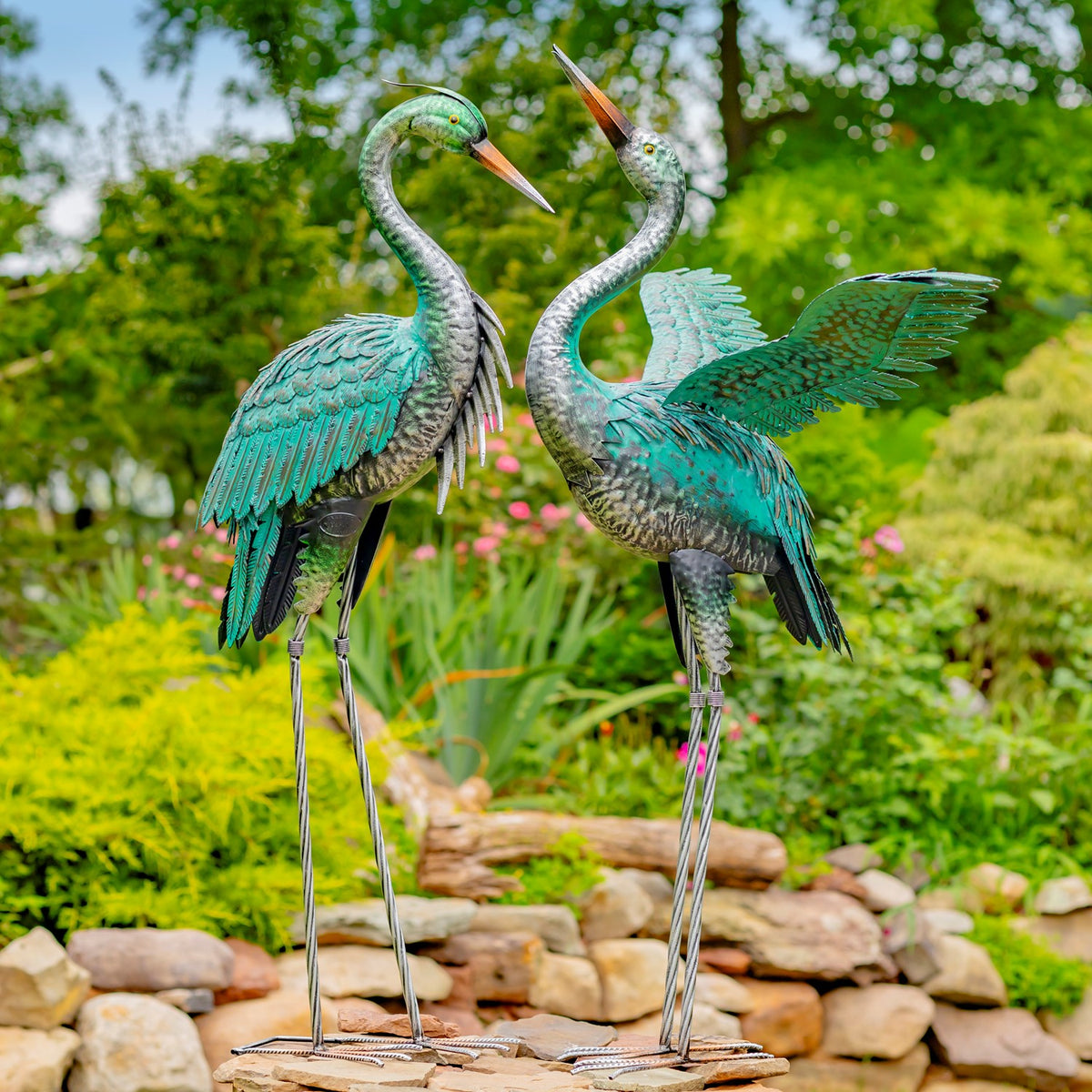 Two teal metal heron sculptures stand together among flowers and stone edging.
