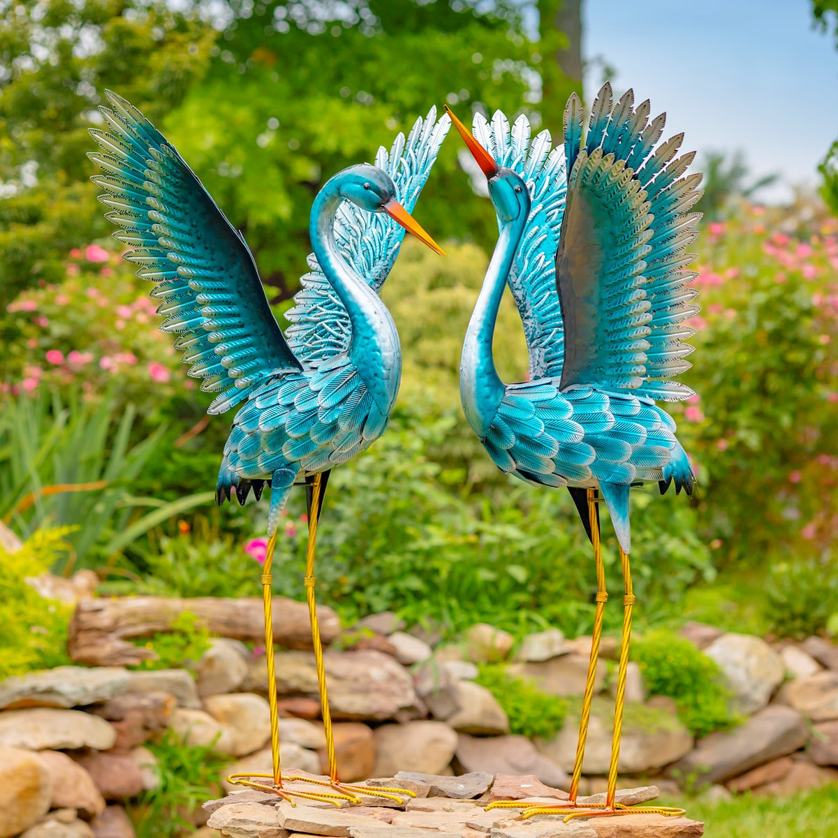 Pair of blue-green crane garden figurines with raised wings beside a stone garden border