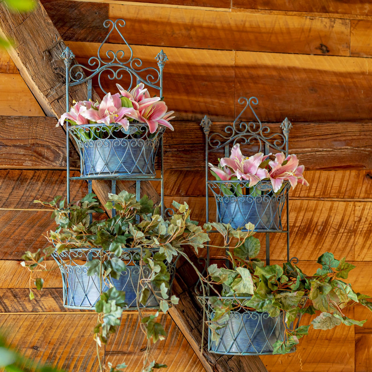 Two ornate blue wall planters filled with pink flowers and trailing greenery against a wooden wall.