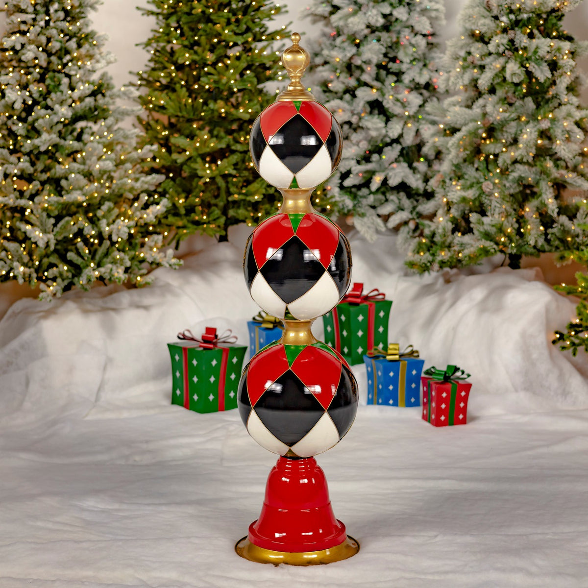 A single tower of three red, black and white patterned ornaments stands on a gold base before snowy Christmas trees.