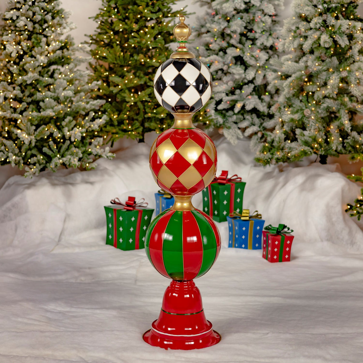 A red-based decorative tower topped with three colorful patterned spheres stands among holiday trees and gifts.