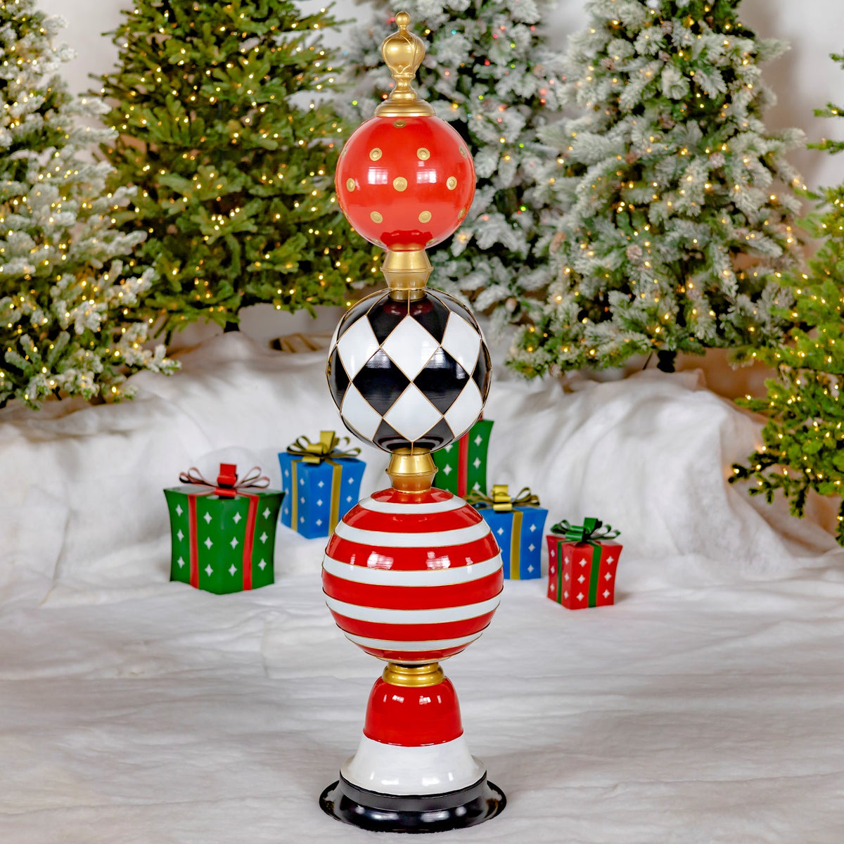 A tall red, black, white, and gold ornament tower stands on a snowy floor before lit Christmas trees.