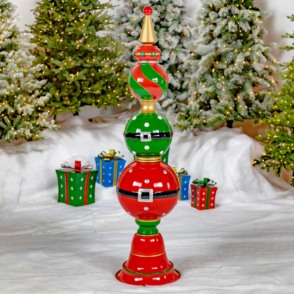 Red and green ornament tower with black belt details stands among wrapped gifts and snowy Christmas trees.