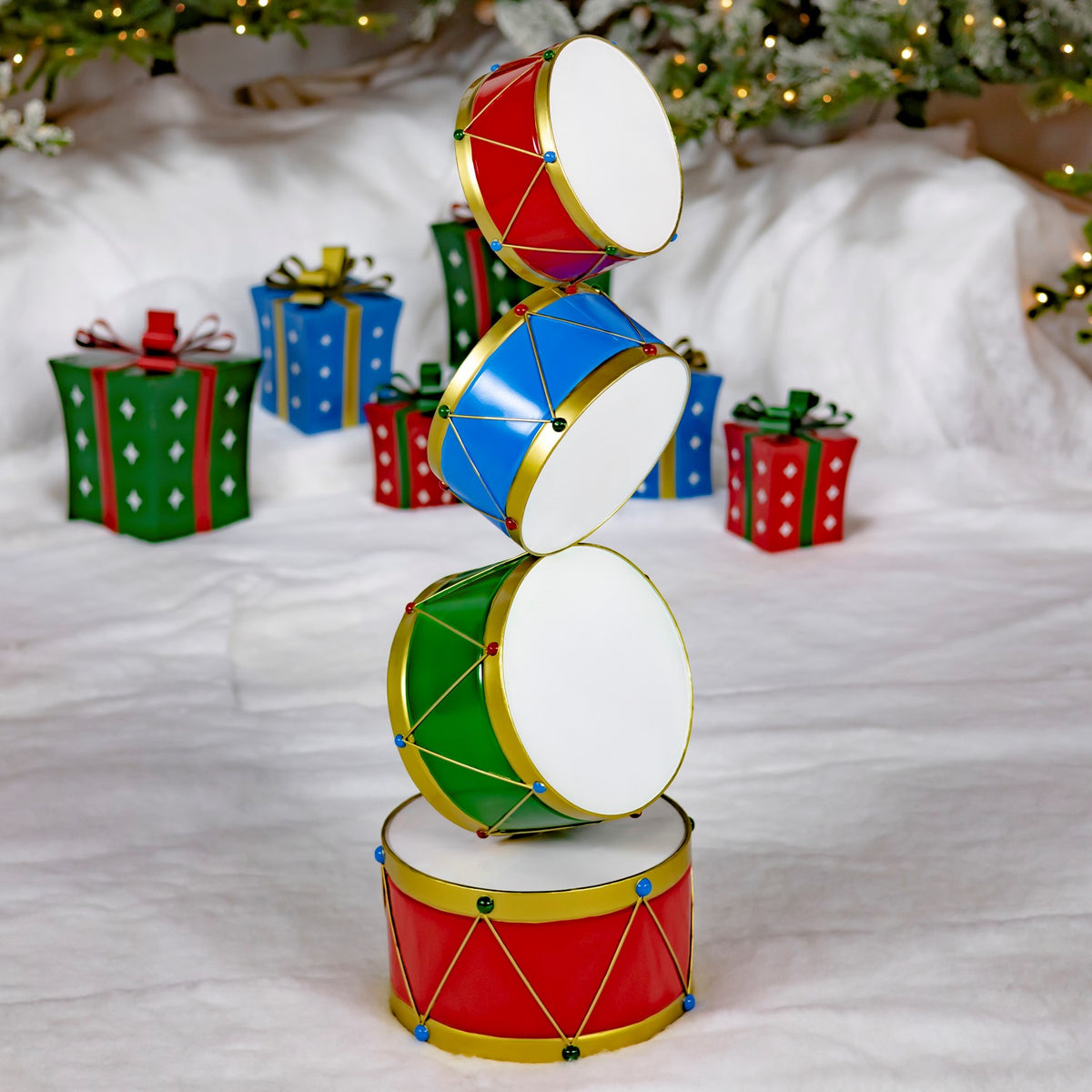 A colorful tower of four red, blue, and green drums stands among Christmas gifts and tree branches.