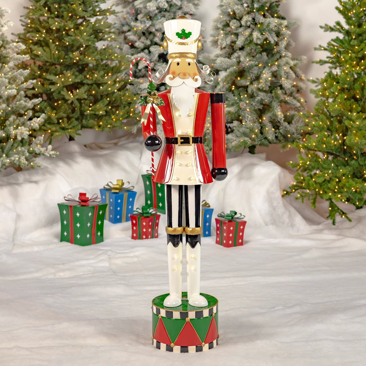 Full-length nutcracker holding a candy cane, standing among snowy trees and wrapped gifts.