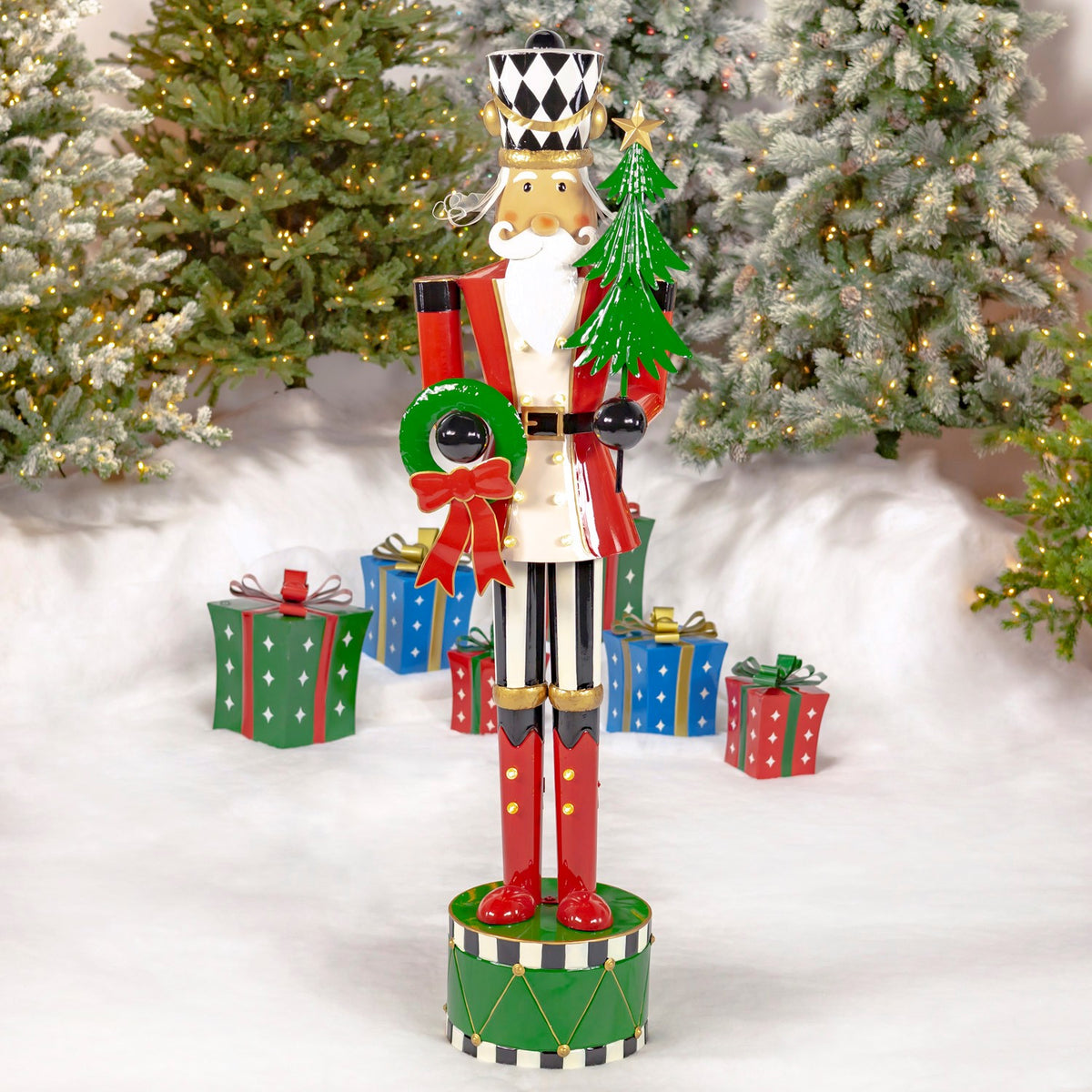 Full-length red-and-white nutcracker holding a green tree and wreath among wrapped gifts and lit Christmas trees.