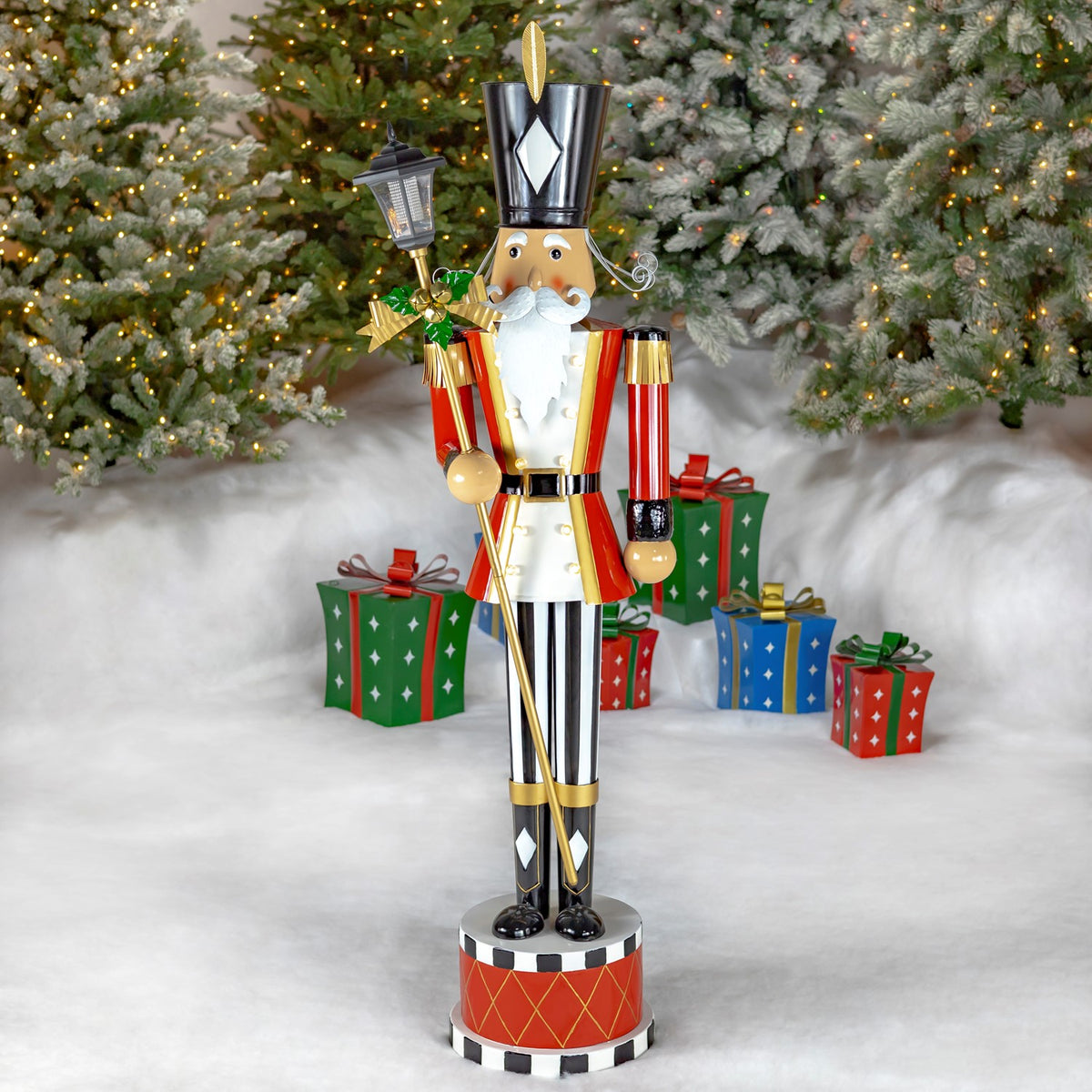 Full view of a red-and-white nutcracker holding a lantern, posed among snowy trees and wrapped gifts.