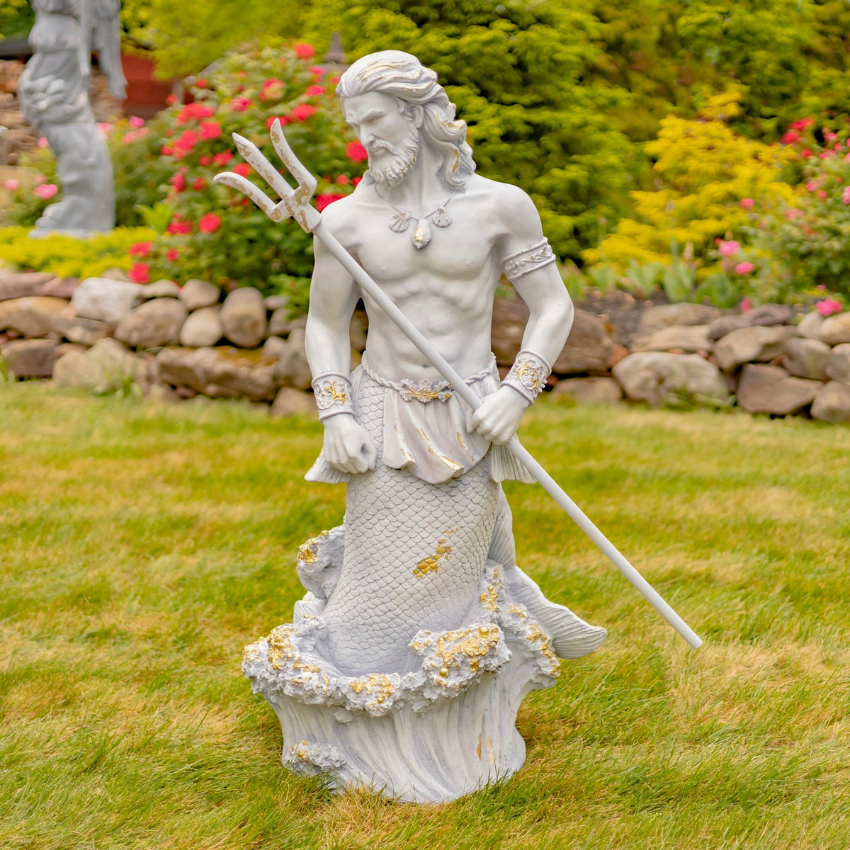 Pale stone-colored merman statue stands on a wave base, holding a trident in a flower-filled garden.