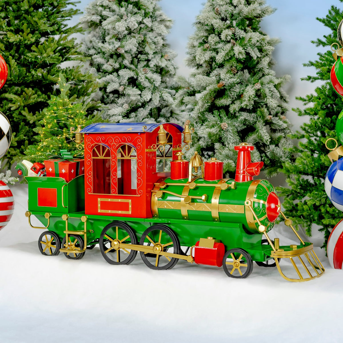 A green and red decorative train with a carriage and gold trim sits on snow among frosted Christmas trees.