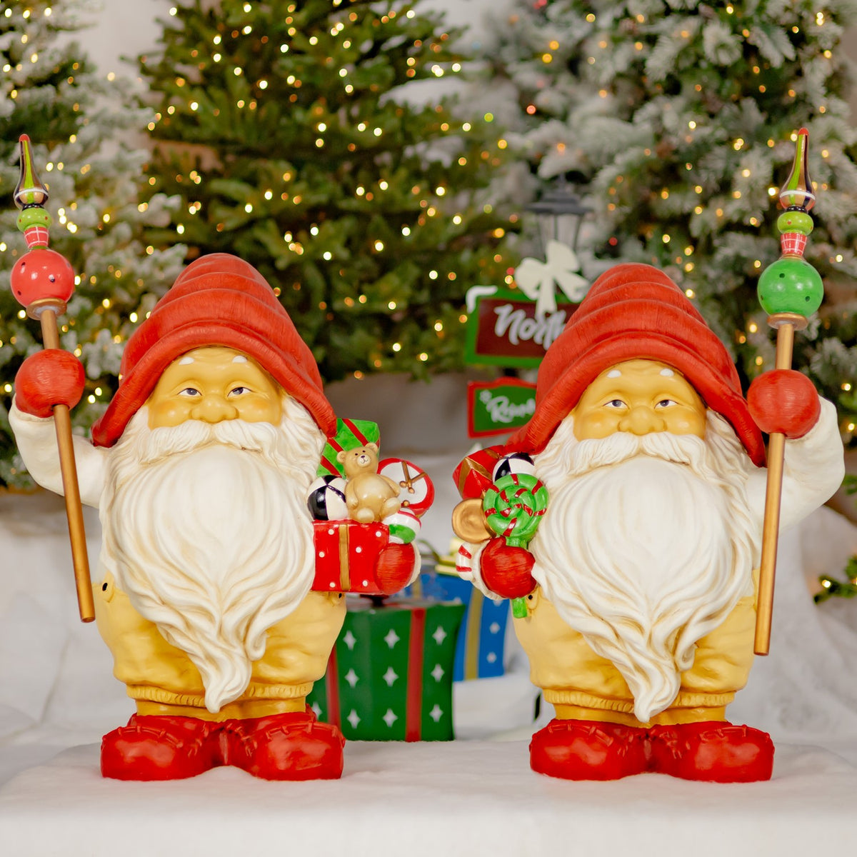 Two red-hatted Santa gnome statues stand with decorated staffs and gifts in a festive winter display.
