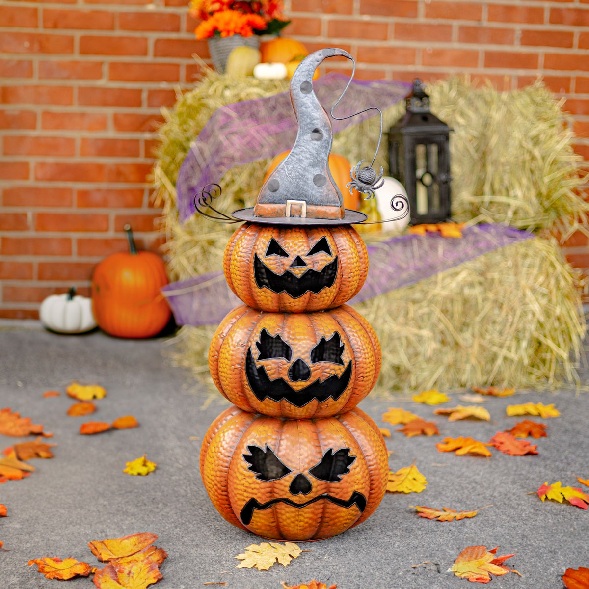 Three stacked orange jack-o’-lanterns wear a crooked gray witch hat, with a dangling spider and autumn leaves nearby.