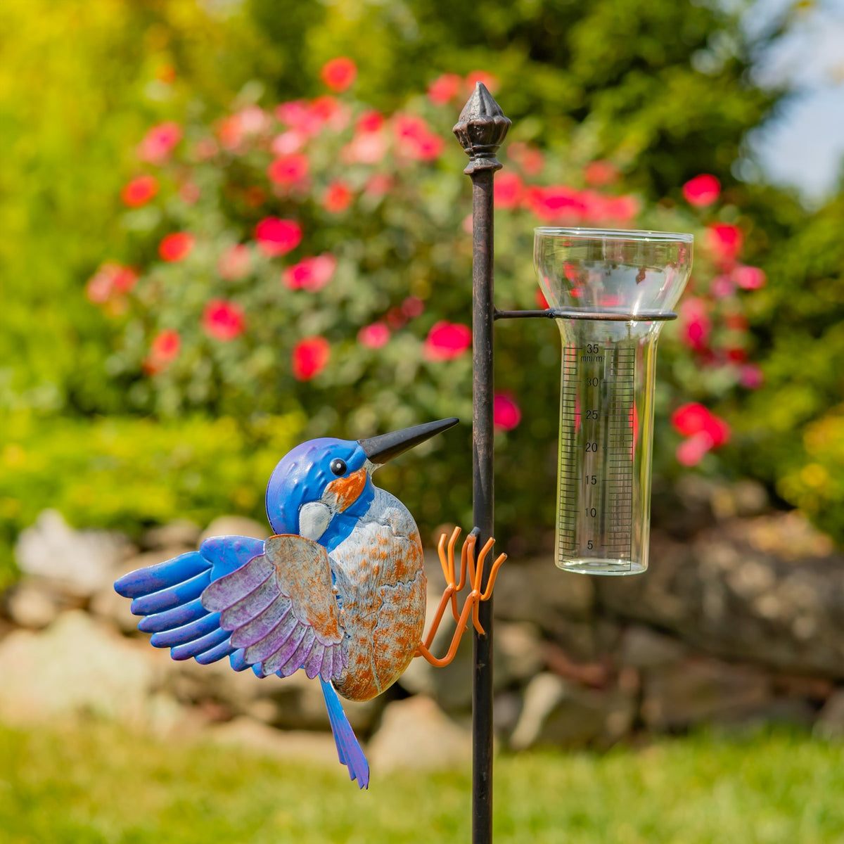Blue and orange hummingbird garden stake beside a glass rain gauge, close view