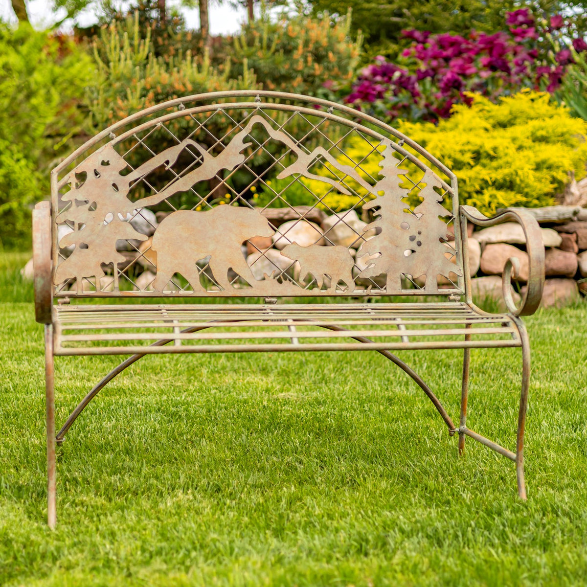 Arched metal garden bench with bear and mountain silhouettes, photographed on a lawn among flowers.