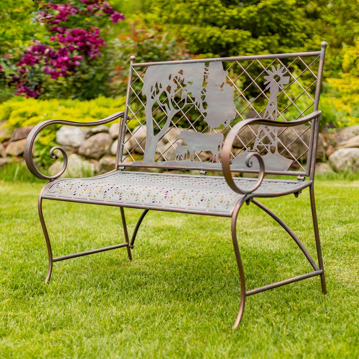 Metal garden bench on grass, with curled arms and a backrest featuring a cow, tree and windmill silhouette.