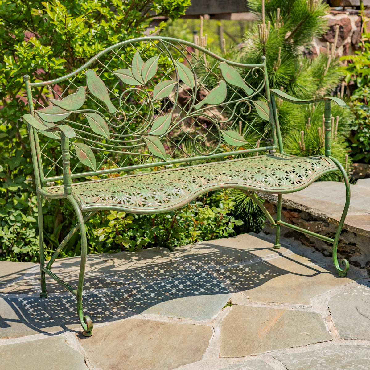 Green metal garden bench with an ornate leaf-patterned backrest, shown among garden greenery.