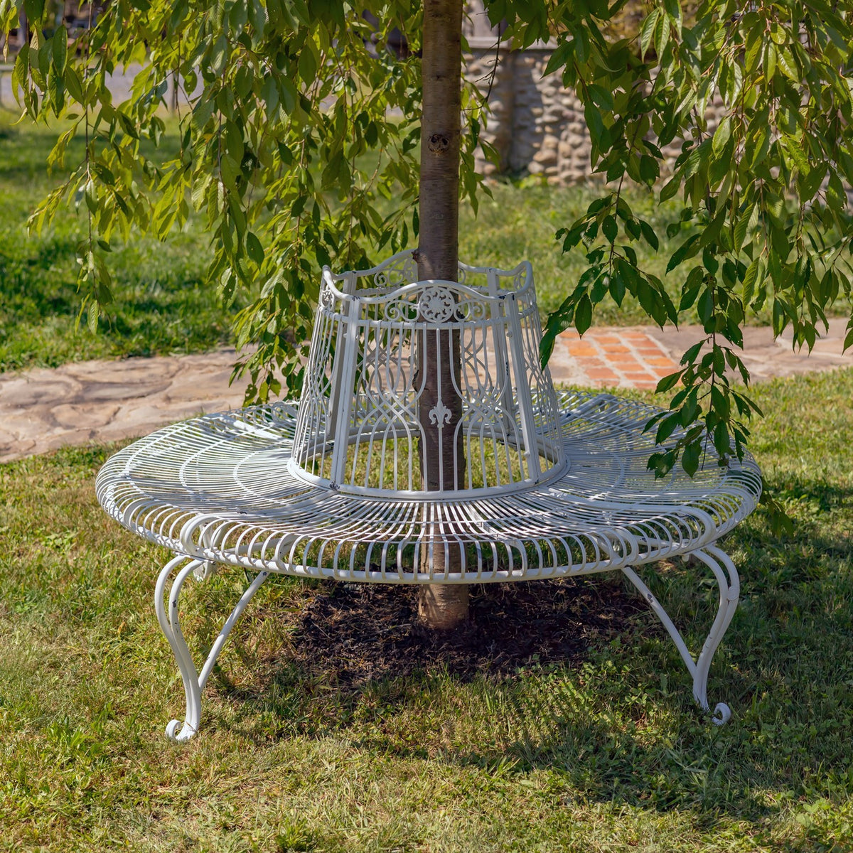 White circular metal bench wraps around a tree in a sunny garden.