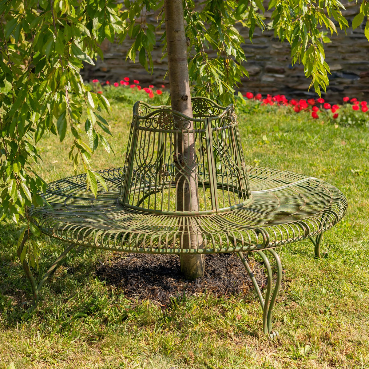 Dark bronze circular bench surrounds a tree amid grass and flowers.