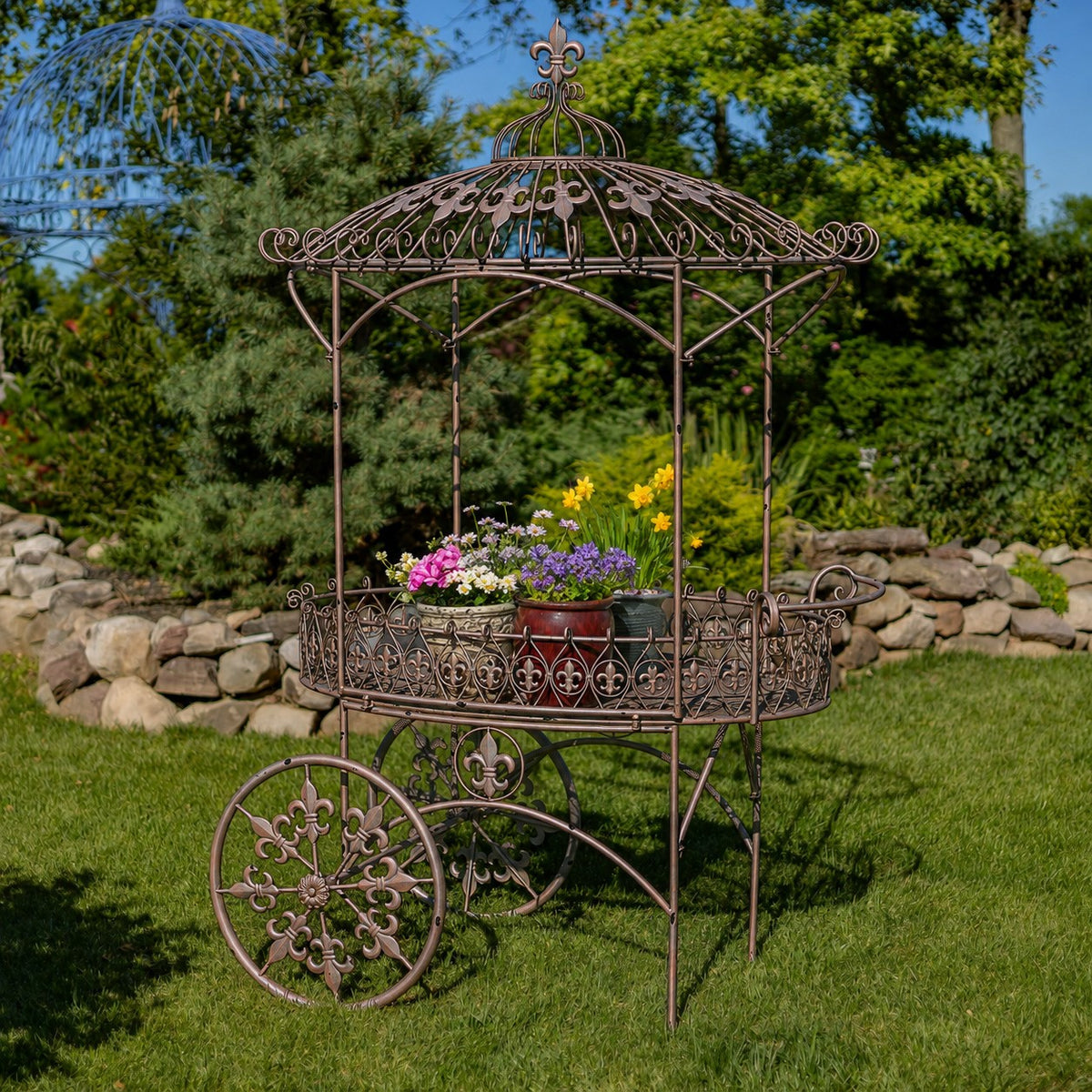 Dark bronze iron flower cart with decorative canopy, large wheels, and colorful potted flowers outdoors.
