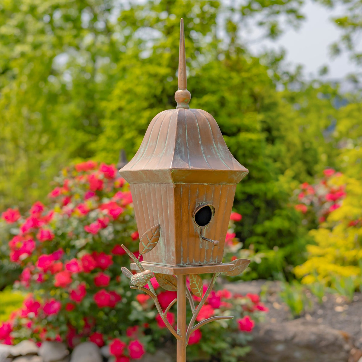 Antique copper birdhouse on a garden stake, framed by bright flowers and greenery.