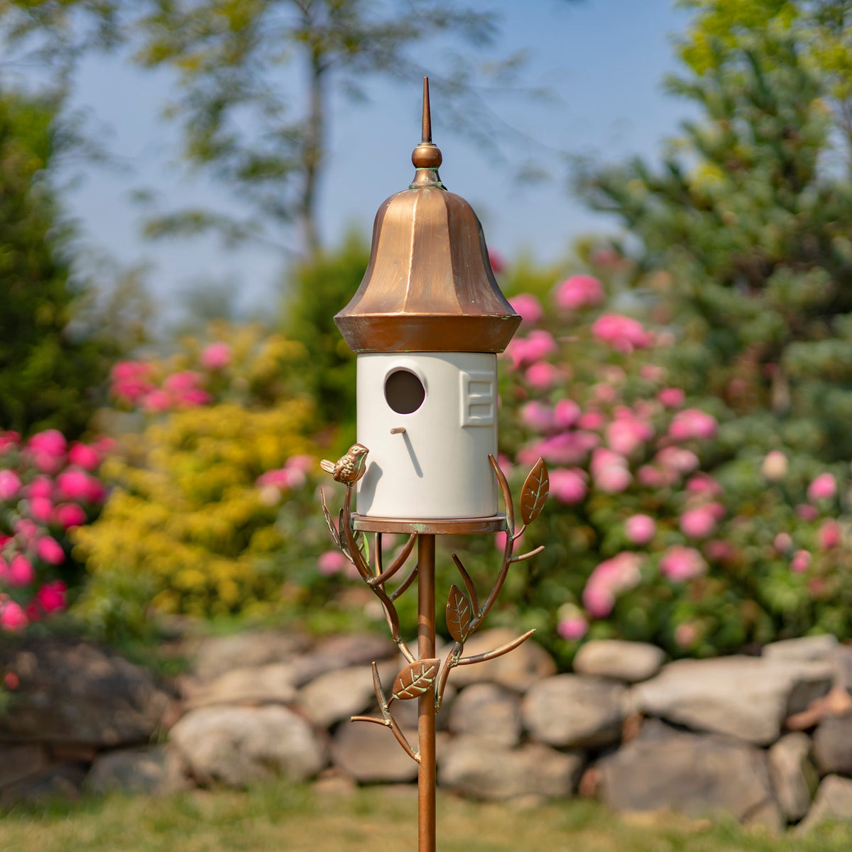 A decorative birdhouse stake with a copper-colored roof and metal branch details stands among flowering garden plants.