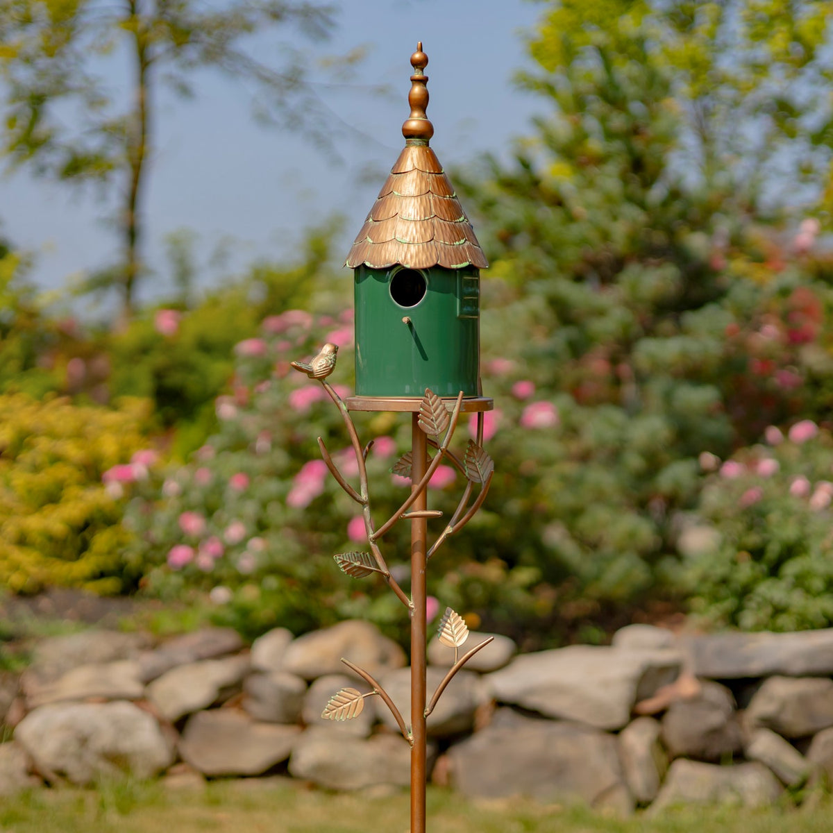 Decorative green birdhouse with a copper-toned roof and leafy metal branches, standing among garden flowers.