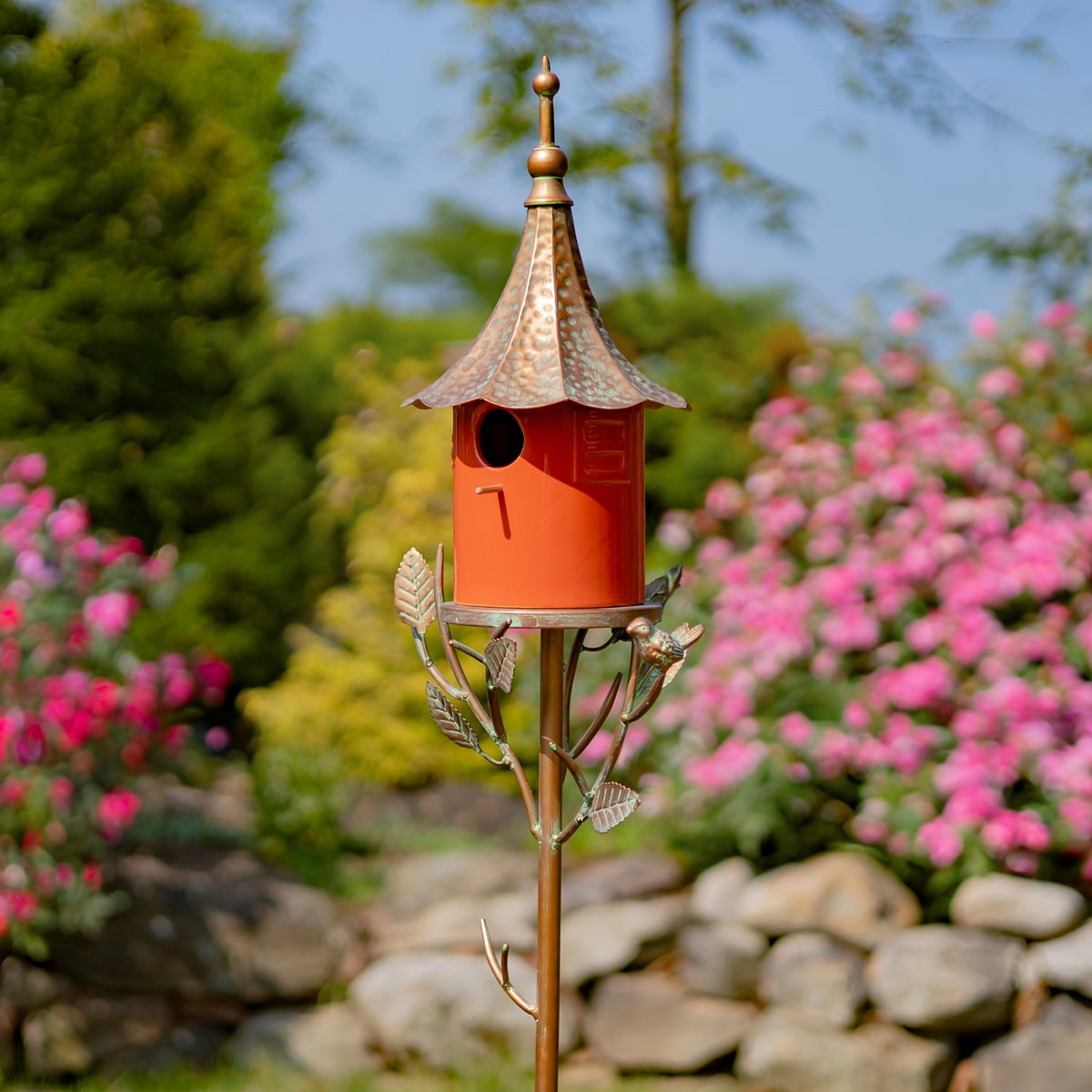 A red birdhouse with a weathered copper-colored roof sits atop a leafy metal garden stake among flowers.