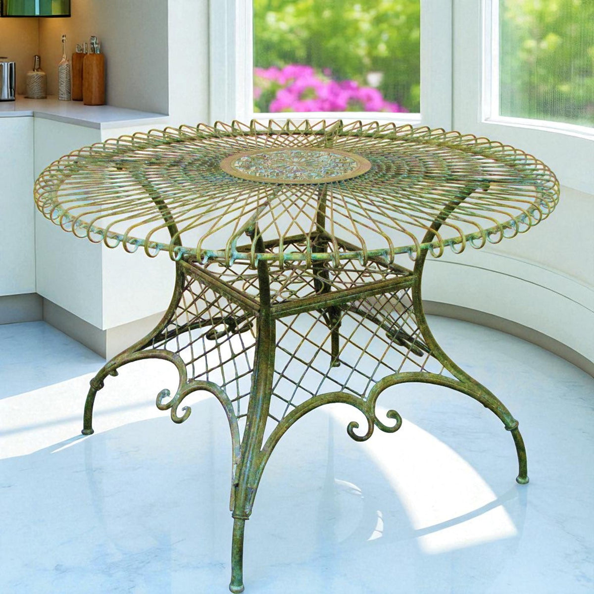 Ornate round green iron table with a woven wire top and curled legs in a bright sunroom.