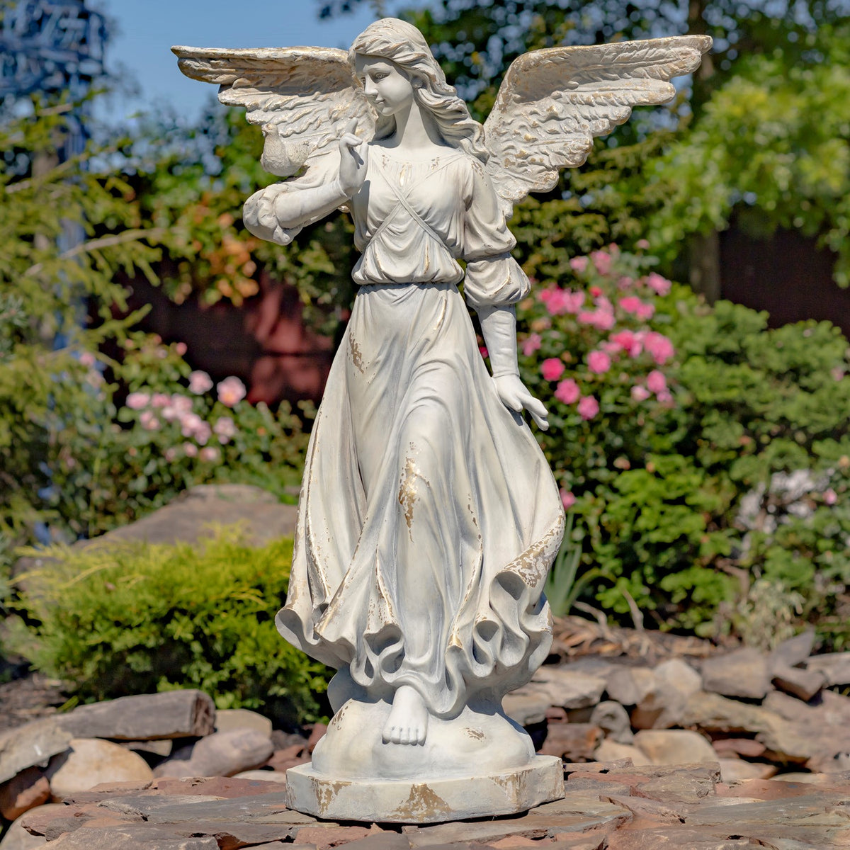 Ivory angel statue with broad wings and a bird on her arm, shown full length in a garden.