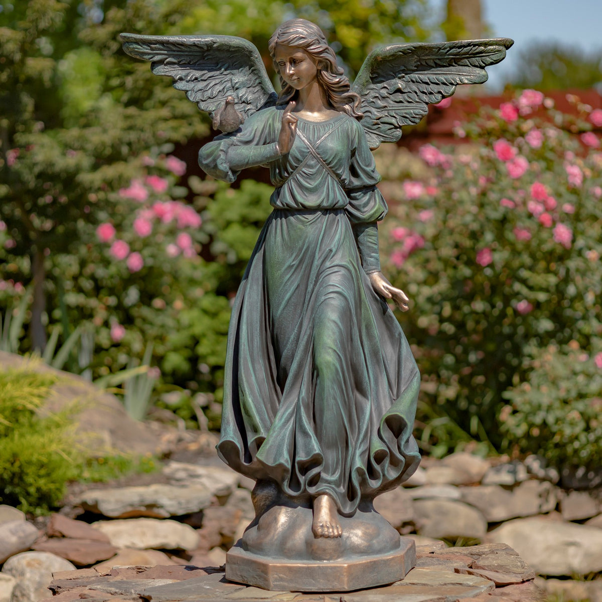 Full-length angel statue in a flowing green dress, with outstretched wings and a small bird, standing among garden stones.