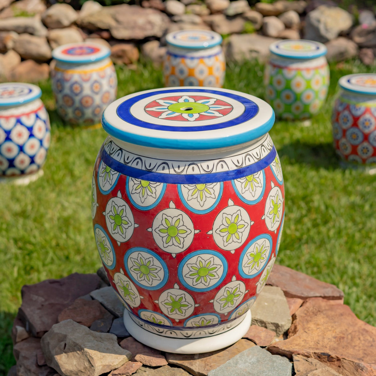 A red floral-patterned ceramic stool sits on rocks in grass, with several differently patterned stools behind it.