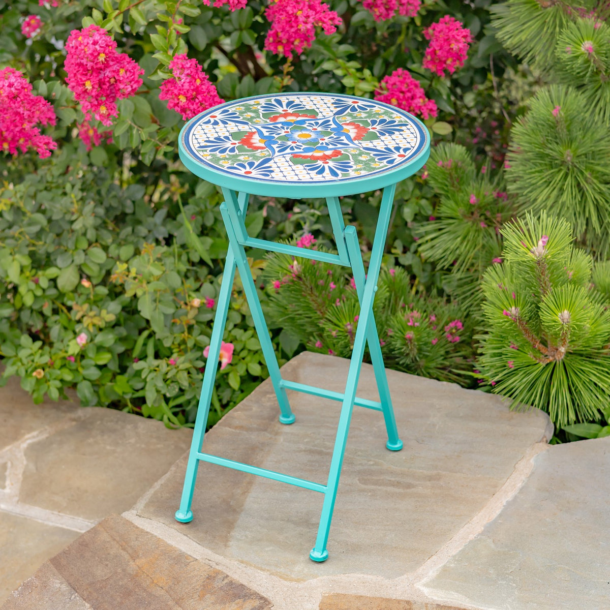 Turquoise folding plant stand with a colorful round mosaic top among pink flowers on a stone patio.