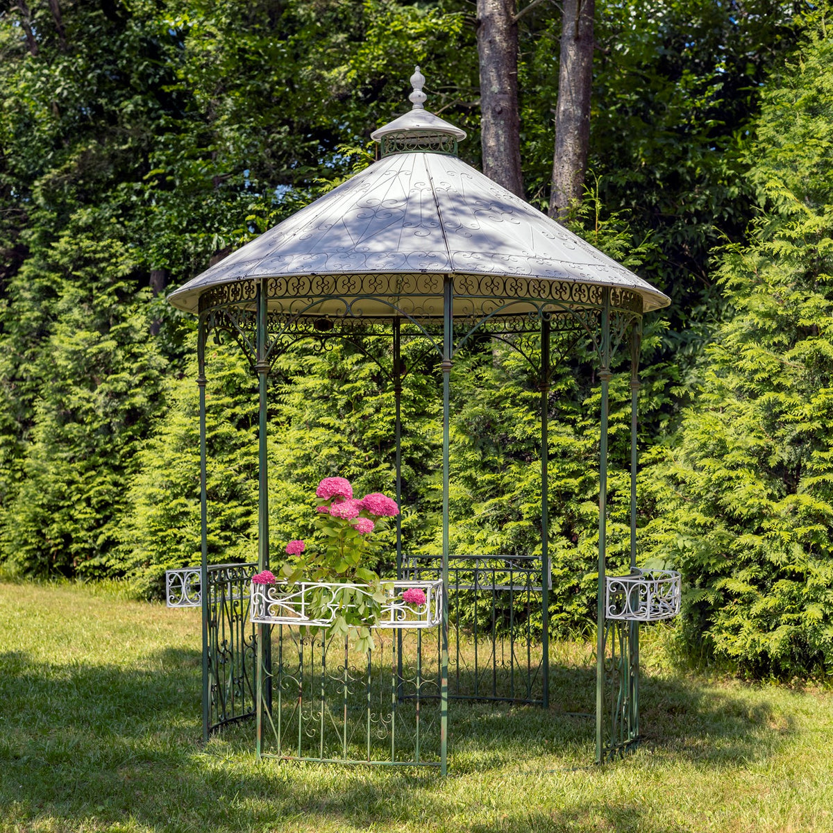 Ornate green metal gazebo with a pale roof and flower baskets set on a lawn among trees.